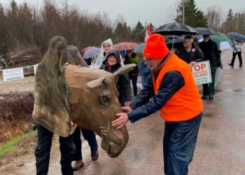 A group of protesters walk down a wet road under umbrellas. In the foreground, a person wears a large, brown, sculpted moose head mascot. A man in an orange safety vest and hat reaches out to pet the moose head.