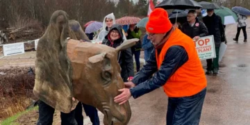 A group of protesters walk down a wet road under umbrellas. In the foreground, a person wears a large, brown, sculpted moose head mascot. A man in an orange safety vest and hat reaches out to pet the moose head.