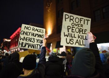 Protesters hold signs reading "No races only humans, where's your humanity" and "Profit over people will kill us all" at a nighttime rally against ICE in Minneapolis.