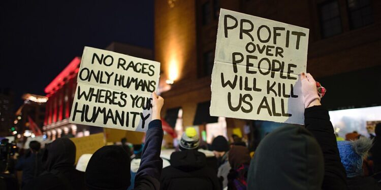 Protesters hold signs reading "No races only humans, where's your humanity" and "Profit over people will kill us all" at a nighttime rally against ICE in Minneapolis.