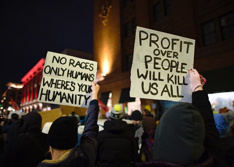 Protesters hold signs reading "No races only humans, where's your humanity" and "Profit over people will kill us all" at a nighttime rally against ICE in Minneapolis.