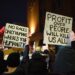 Protesters hold signs reading "No races only humans, where's your humanity" and "Profit over people will kill us all" at a nighttime rally against ICE in Minneapolis.