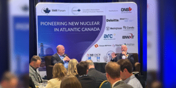 Two men sit on stage holding microphones during a panel discussion, in front of a backdrop reading 'Pioneering New Nuclear in Atlantic Canada' with sponsor logos including Westinghouse, NB Power, NWMO, and Candu. An audience is seated in the foreground.