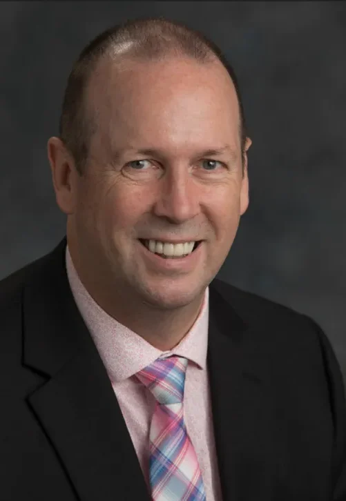 Headshot of New Brunswick Auditor-General Paul Martin, wearing a black blazer and plaid tie, smiling against a dark grey background.