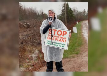 A person wearing a clear rain poncho holds a megaphone and a sign reading 'Stop the Tantramar Gas Plant — Clean Air, Clean Water, Clean Energy for All,' standing on a dirt road in overcast, rainy conditions.