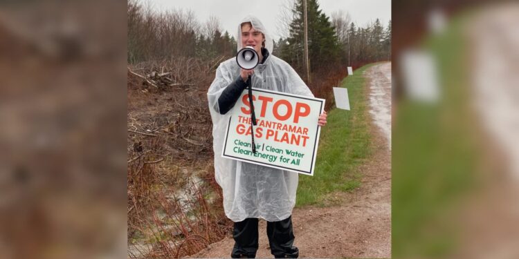 A person wearing a clear rain poncho holds a megaphone and a sign reading 'Stop the Tantramar Gas Plant — Clean Air, Clean Water, Clean Energy for All,' standing on a dirt road in overcast, rainy conditions.
