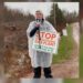 A person wearing a clear rain poncho holds a megaphone and a sign reading 'Stop the Tantramar Gas Plant — Clean Air, Clean Water, Clean Energy for All,' standing on a dirt road in overcast, rainy conditions.