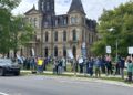 A large crowd of people holding protest signs stands on a lawn in front of a large stone government building on a cloudy day.