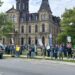 A large crowd of people holding protest signs stands on a lawn in front of a large stone government building on a cloudy day.