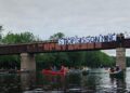 Paddlers pass beneath a bridge bearing a "Stop Sisson Mine" banner and a Wolastoq flag on the Nashwaak River.