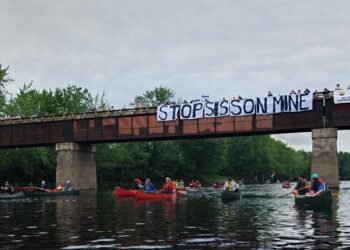 Paddlers pass beneath a bridge bearing a "Stop Sisson Mine" banner and a Wolastoq flag on the Nashwaak River.