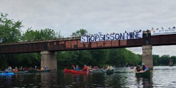 Paddlers pass beneath a bridge bearing a "Stop Sisson Mine" banner and a Wolastoq flag on the Nashwaak River.