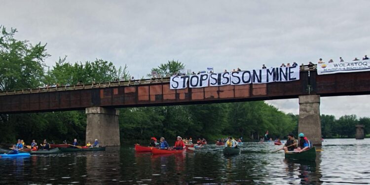 Paddlers pass beneath a bridge bearing a "Stop Sisson Mine" banner and a Wolastoq flag on the Nashwaak River.