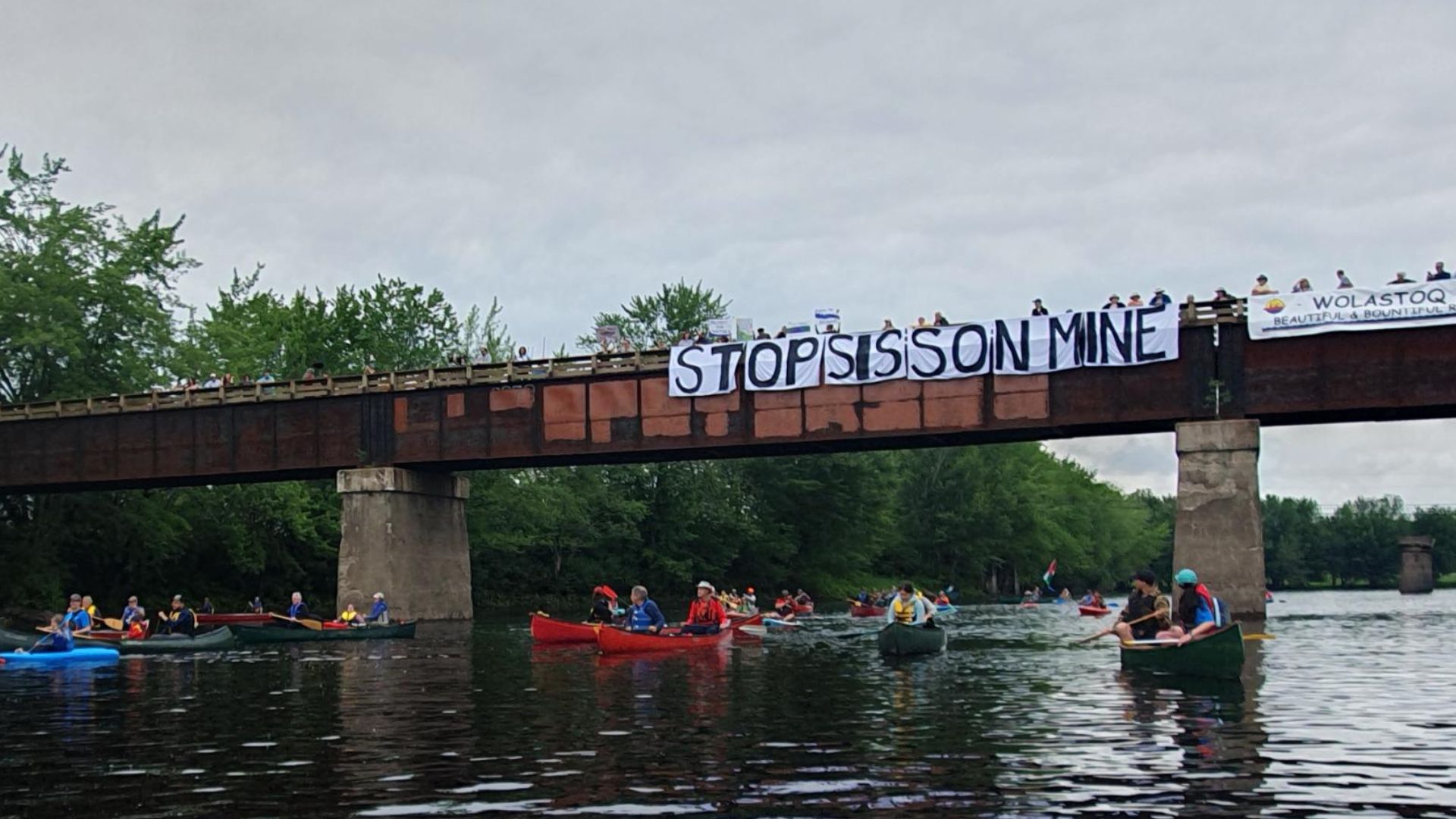 8 Paddlers pass beneath a bridge bearing a "Stop Sisson Mine" banner and a Wolastoq flag on the Nashwaak River.
