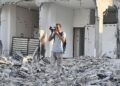 A man in a light shirt and grey pants stands amid the rubble of a destroyed multi-story building, raising a camera to photograph the scene.