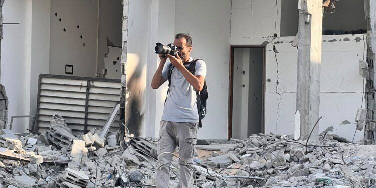A man in a light shirt and grey pants stands amid the rubble of a destroyed multi-story building, raising a camera to photograph the scene.
