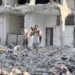 A man in a light shirt and grey pants stands amid the rubble of a destroyed multi-story building, raising a camera to photograph the scene.