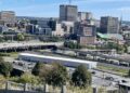 Elevated landscape view of Saint John, New Brunswick, showing an overpass and parking lot in the foreground with mid-rise and high-rise commercial buildings under a clear blue sky.