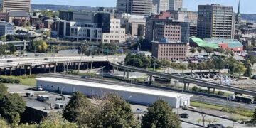 Elevated landscape view of Saint John, New Brunswick, showing an overpass and parking lot in the foreground with mid-rise and high-rise commercial buildings under a clear blue sky.