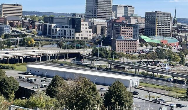 Elevated landscape view of Saint John, New Brunswick, showing an overpass and parking lot in the foreground with mid-rise and high-rise commercial buildings under a clear blue sky.
