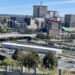 Elevated landscape view of Saint John, New Brunswick, showing an overpass and parking lot in the foreground with mid-rise and high-rise commercial buildings under a clear blue sky.