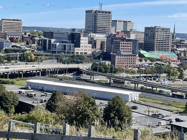 Elevated landscape view of Saint John, New Brunswick, showing an overpass and parking lot in the foreground with mid-rise and high-rise commercial buildings under a clear blue sky.
